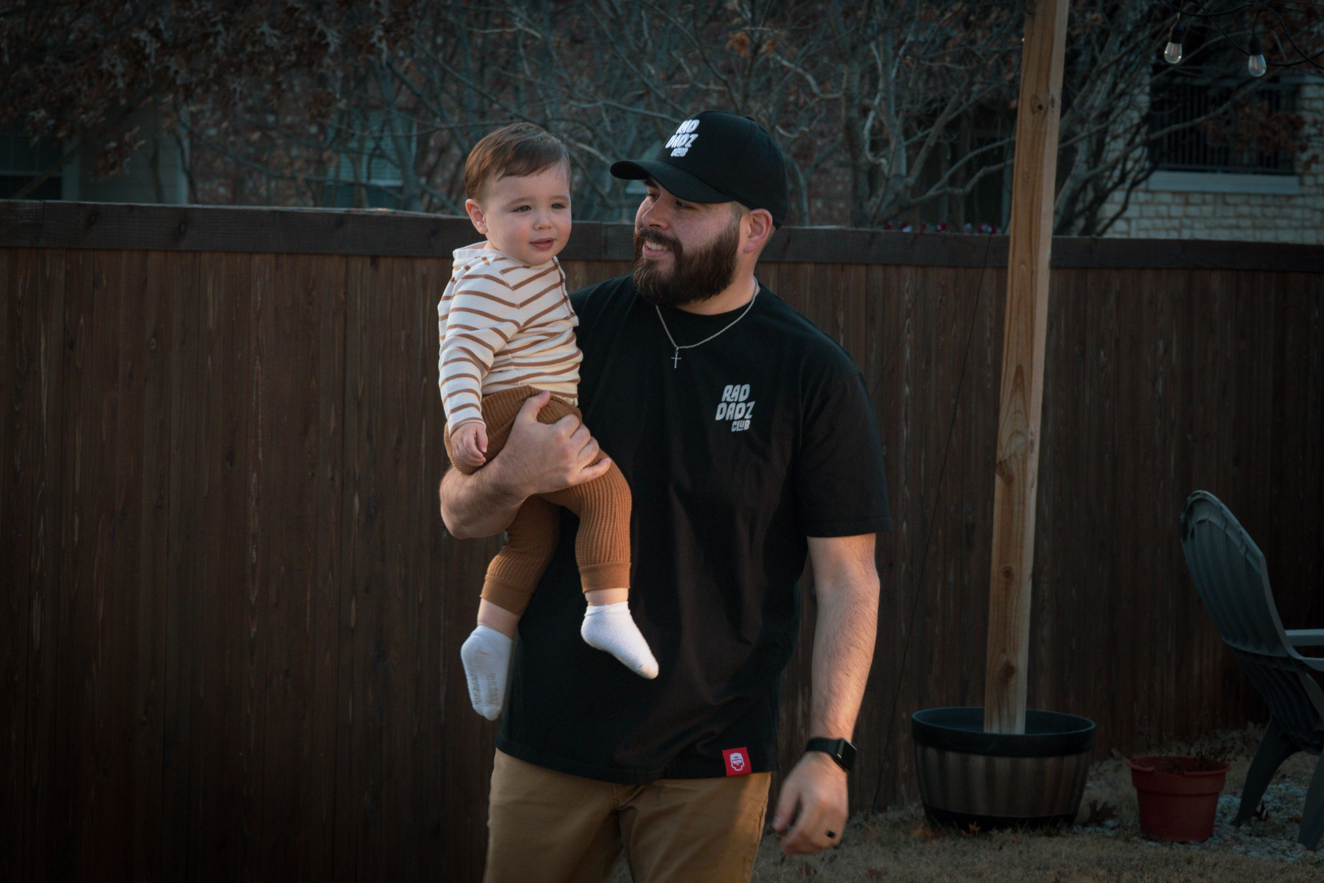 A dad wearing a black cotton shirt with a simple white logo while carrying his son